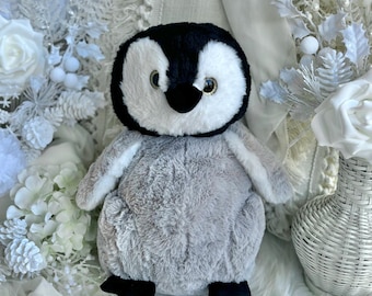 May include: A plush penguin toy with a black head, white face, and gray body. The penguin has black feet and small, light-colored eyes. It is set against a backdrop of white flowers and a fluffy white surface.