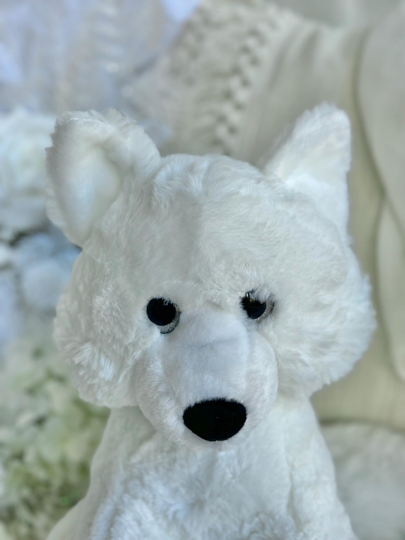 K&ouml;nnte beinhalten: Ein wei&szlig;es Pl&uuml;sch-Eisb&auml;r-Spielzeug mit schwarzen Augen und einer schwarzen Nase. Das Stofftier hat eine flauschige Textur und aufrechte Ohren. Der B&auml;r ist vor einem verschwommenen wei&szlig;en Hintergrund platziert.