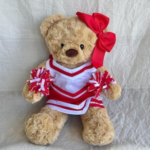 May include: A plush teddy bear dressed as a cheerleader. The bear wears a red and white uniform with matching pom-poms and a large red bow. The bear is light brown with a dark brown nose and black eyes.