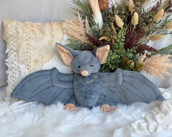 May include: A plush bat toy featuring soft grey fabric, large pointed ears with tan inner lining, a small tan nose, and wide outstretched wings. The bat has a textured grey collar around its neck and small tan feet at the base of its body.