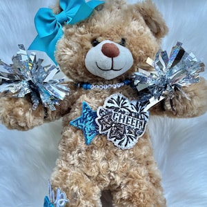 May include: A tan plush teddy bear wearing a bright turquoise hair bow, a beaded necklace spelling PANTHERS, and holding two silver metallic pom-poms. A blue sequined star patch and a white patch with the word CHEER are attached to the chest.