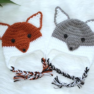 May include: Two crocheted fox hats. One is orange and white, the other is gray and white. Both have fox faces with button eyes and braided ties. The hats are on a white, fluffy surface.