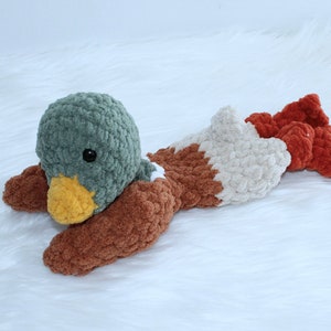 May include: A crocheted plush toy duck with a green head, brown body, and white and orange accents. The duck is lying on its belly with its head turned to the side.