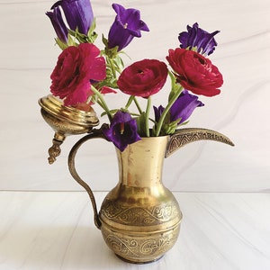 May include: A vintage brass pitcher with intricate etched designs, holding a vibrant arrangement of red ranunculus and purple bellflowers. The pitcher has a curved spout and a handle, creating a striking contrast with the colorful flowers.