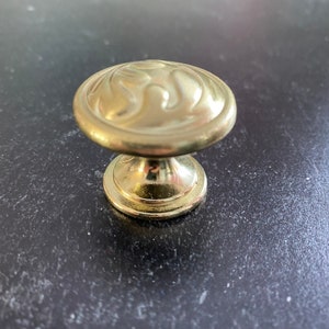 May include: A round, gold-colored metal cabinet knob featuring a smooth, raised, swirling embossed pattern on its top surface. The knob has a polished finish and a circular base that tapers upward to support the rounded, decorative head.