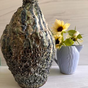 May include: A textured, handcrafted ceramic vase with a rough, organic appearance. The vase features a blend of earthy tones, including brown, green, and blue. A small, light blue pitcher holds yellow sunflowers. Home decor.