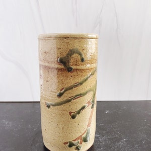 May include: A cylindrical ceramic vase with a speckled tan and brown glaze. The vase features a tree branch design in green and brown, adding a natural aesthetic. The vase is approximately 6 inches tall.