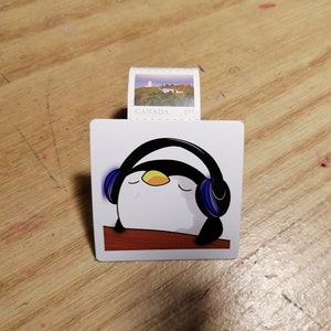 Peut inclure: Un pingouin blanc aux yeux noirs et au bec jaune portant un casque audio bleu. Le pingouin est sur un fond brun avec le texte "CANADA 19¢" au-dessus.