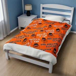 Può includere: Una coperta in pile arancione e nera con un motivo ripetitivo della mascotte dei Philadelphia Flyers, Gritty. La coperta presenta il testo "Gritty's #1 Fan" e "Philadelphia Flyers".