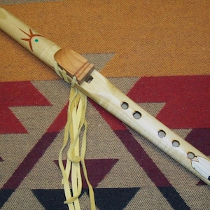 May include: A light-colored wooden Native American style flute featuring a carved block, five finger holes, and decorative painted symbols including a sun and a feather. The instrument is adorned with long, thin, light yellow leather fringe ties.