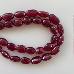 Puede incluir: Una hebra de cuentas de piedras preciosas ovaladas de color rojo oscuro. Las cuentas son lisas y pulidas, con un acabado brillante.