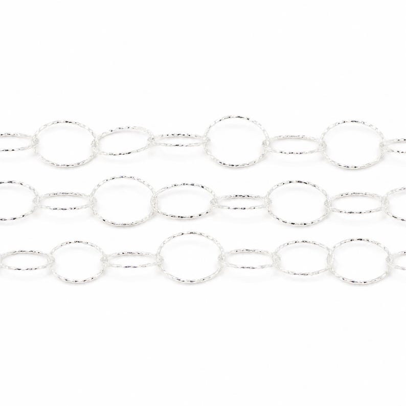May include: A silver-toned metal chain featuring a repeating pattern of alternating large and small circular links. Each individual link has a textured, shimmering surface that reflects light, creating a consistent metallic appearance throughout.