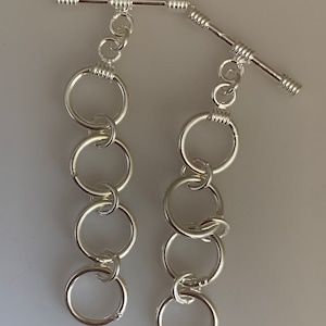 May include: Two silver toggle clasps with a chain of five silver rings. The clasps are made of a straight bar with a twisted design.