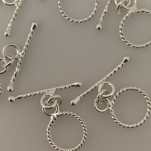 Könnte beinhalten: Silberne Toggle-Verschlüsse mit einem gedrehten Seil-Design. Die Verschlüsse haben einen runden Ring an einem Ende und eine Stange mit einem runden Ring am anderen Ende.