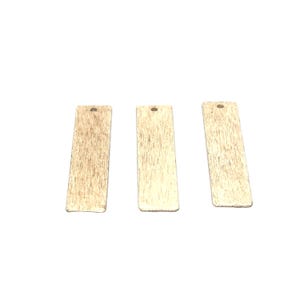May include: Three rectangular, light-colored wooden pendants with a textured surface. Each pendant has a small hole at the top for stringing. The pendants are arranged against a white background.
