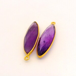 May include: Two elongated, faceted amethyst gemstones with a deep purple hue, set in gold-colored bezels. Each gemstone has a small loop at one end, suggesting they are designed for jewelry making.