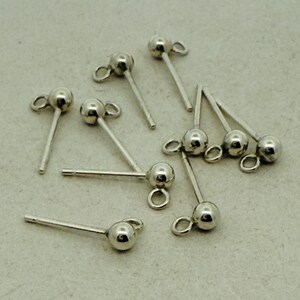 May include: A selection of small, silver-coloured earring posts. Each post features a ball at one end and a loop for attaching a charm. The posts are scattered on a light grey surface. The earrings are minimalist.