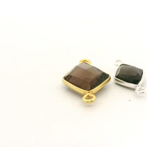 May include: Two gemstone pendants. One is a square-shaped, smoky quartz gemstone with a gold-colored bezel and two loops. The other is a rectangular, smoky quartz gemstone with a silver-colored bezel and two loops. Both are on a white background.