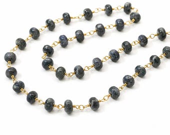 May include: A necklace featuring a series of round, dark gray, speckled beads linked together by thin gold-toned wire. The beads are spaced evenly along the chain, which is arranged in a loose, curved formation against a plain white background.