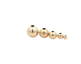 May include: Five gold-colored, spherical beads of varying sizes are arranged in a diagonal line. Each bead has a small, dark hole at the top. The beads are smooth and reflective, with a warm, metallic sheen. The smallest bead is approximately 0.25 inches in diameter.
