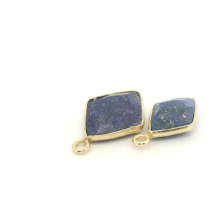 May include: Two blue gemstone pendants with gold-colored bezels. The gemstones are diamond-shaped with a textured surface. Each pendant has a small loop at the bottom for attaching to a chain or other jewelry component.