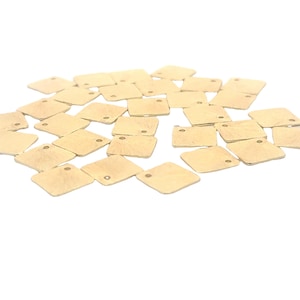 May include: A pile of gold-colored metal square charms with a small hole in each corner. The charms are scattered on a white background.