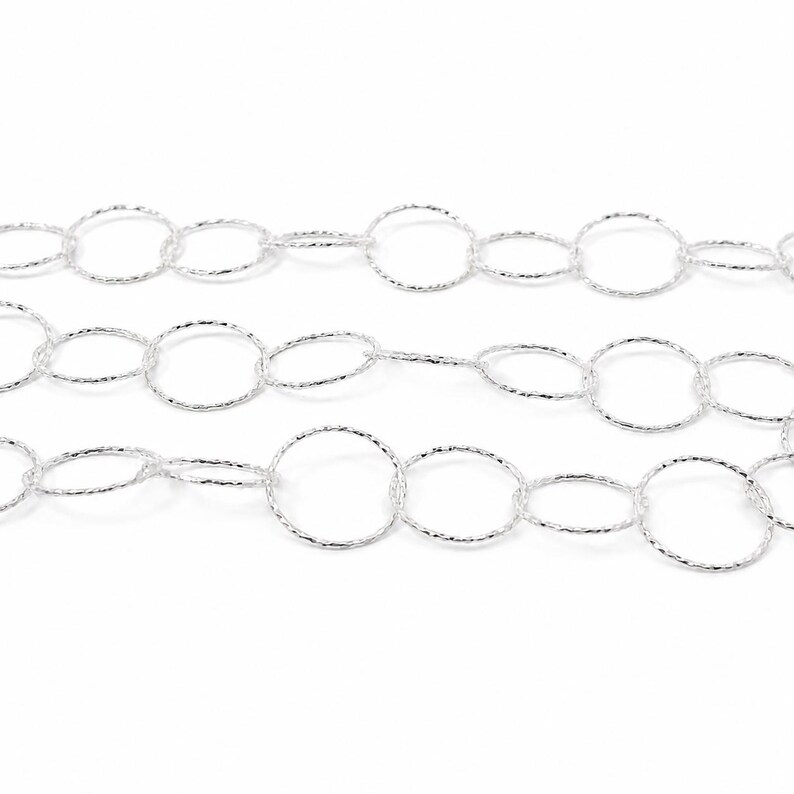 May include: A silver-toned metal chain composed of interconnected circular links. Each individual link features a textured, faceted surface that reflects light, creating a shimmering appearance across the entire length of the jewelry chain.