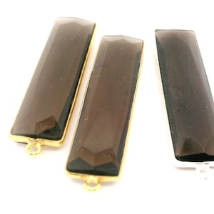 May include: Three rectangular smoky quartz gemstone pendants. Two are set in gold-colored metal, and one in silver-colored metal. Each has a faceted surface and a loop for attaching to jewelry. The background is white.