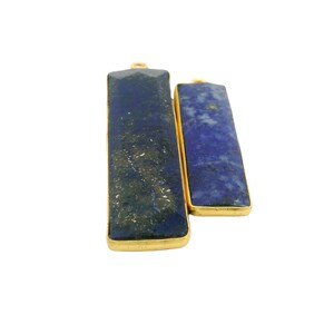 May include: Two rectangular pendants with gold-colored frames. The pendants feature a dark blue stone with gold flecks and a lighter blue stone. The pendants are displayed against a white background.