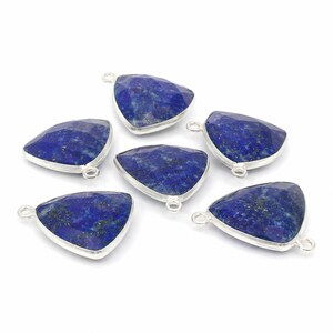 Puede incluir: Seis dijes de piedra preciosa lapislázuli de forma triangular engastados en marcos de metal plateado. Cada pieza presenta una superficie facetada de color azul intenso con motas doradas y un pequeño aro circular en la parte superior.