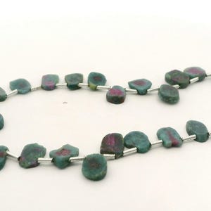 May include: Strands of natural gemstone beads. The beads are a mix of teal and magenta colors, with irregular shapes. The beads are strung on a silver-colored wire. The background is plain white.