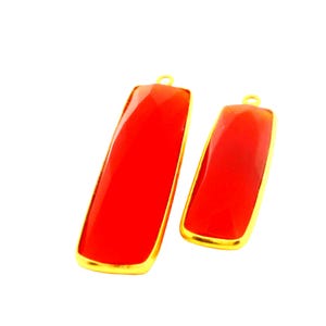 Peut inclure: Deux pendentifs rectangulaires en pierres précieuses rouges avec des bordures en métal doré. Les pendentifs sont de tailles différentes et ont une petite boucle en haut pour les suspendre. Les pierres ont une surface facettée.