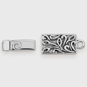 May include: A silver-toned rectangular jewelry clasp consisting of two pieces. One piece features a smooth, polished surface with a circular indentation, while the other displays an intricate, raised scrollwork pattern and a small circular loop.