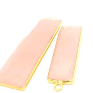 May include: Two rectangular pendants with a light pink center and gold-colored trim. One pendant is longer than the other. The smaller pendant has a gold-colored loop at the bottom. The background is white.