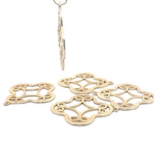 May include: Five gold-colored metal charms with a decorative, intricate, openwork design. The charms are shaped like a four-leaf clover.