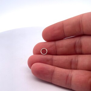 May include: A small, silver jump ring, approximately 5mm in diameter, is held between two fingers.