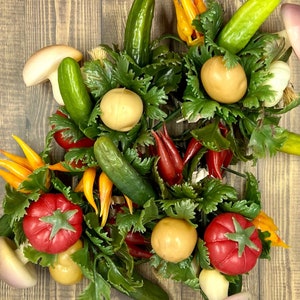 May include: An overhead shot of a collection of artificial vegetables, including cucumbers, tomatoes, peppers, and mushrooms, arranged with green leafy accents. The vegetables are in various colors, including red, yellow, and green, and are set against a wooden background.