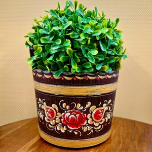 May include: A decorative, dark brown wooden planter with a vibrant green plant. The planter features a hand-painted floral design in red, gold, and white. The rim is accented with gold and red details, creating a rustic and charming aesthetic.