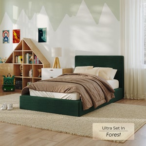 May include: A green upholstered bed frame with a brown blanket and two white pillows. The bed is on a beige rug in a room with a wooden floor and a white wall with a green mountain design.