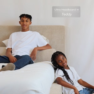 May include: Two people are sitting on a bed with a white headboard. The headboard has the text "Ultra Headboard In Teddy" on it. The person on the left is wearing a white t-shirt and blue jeans. The person on the right is wearing a white t-shirt and blue jeans.