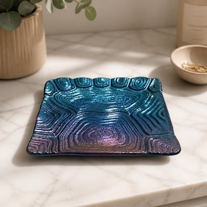 May include: A square glass tray featuring a deep blue and purple iridescent finish with a textured, concentric ripple pattern across the surface. The edges of the tray are slightly scalloped, and the material has a reflective, metallic sheen.