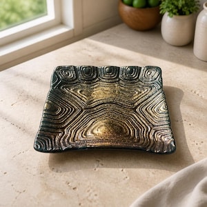 May include: A square glass dish featuring a textured surface with a raised, repeating pattern that resembles a turtle shell. The dish displays a gradient color scheme, transitioning from a deep, dark green at the edges to a metallic gold center.