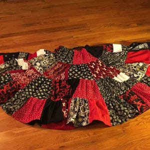 Puede incluir: Una falda patchwork en forma semicircular, con una variedad de telas estampadas. La falda incorpora telas rojas, negras y blancas con diseños de cachemira, lunares y geométricos. La falda está colocada sobre un suelo de madera.
