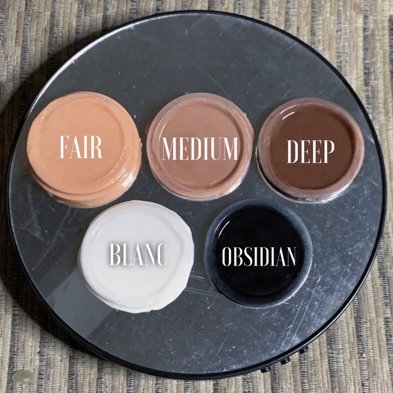 May include: Six small round containers with different shades of foundation makeup. The containers are labeled "Fair", "Medium", "Deep", "Blanc", and "Obsidian".