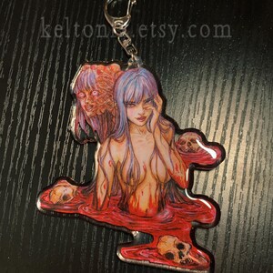 May include: A clear acrylic keychain featuring a woman with long purple hair, a skull, and red liquid. The woman is partially submerged in the liquid, and there are skulls floating around her. The keychain has a silver metal ring.
