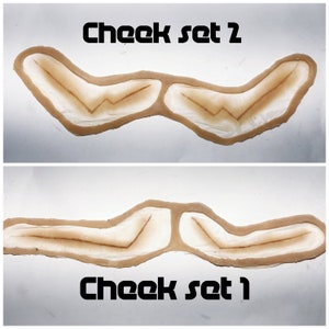 May include: Two sets of brown cheek prosthetics. The top set is labeled "Cheek set 2" and the bottom set is labeled "Cheek set 1".