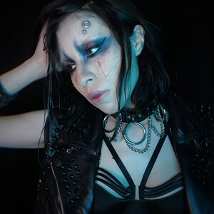 May include: A woman with dark hair and blue and white face paint wears a black leather jacket with studs, a black bra with straps, and a silver chain choker with a large ring.