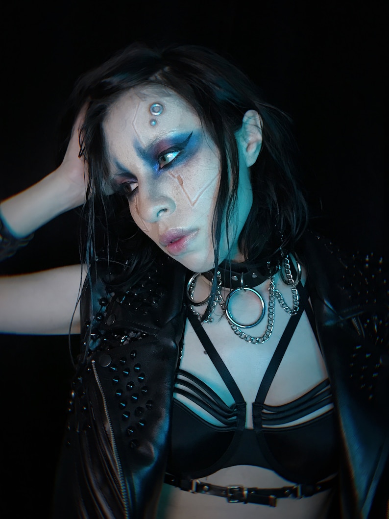 May include: A woman with dark hair and blue and white face paint wears a black leather jacket with studs, a black bra with straps, and a silver chain choker with a large ring.