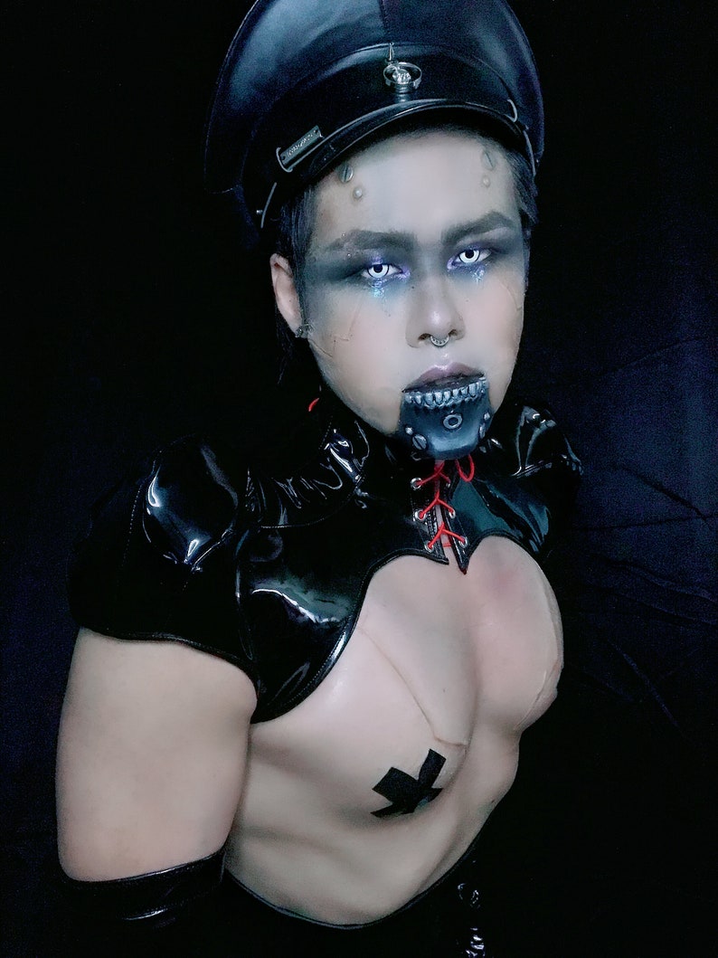 May include: A person wearing a black leather outfit with a black cap, a black choker with red laces, and a black and white cross on their chest.