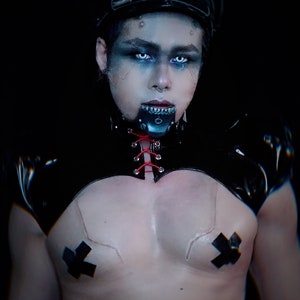 May include: A person wearing a black leather choker with red laces, a black leather top with a lace-up front, and a black cap. They have blue eyeshadow and white contact lenses. They are wearing black tape over their chest.